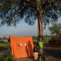 Kvinde og barn foran et stort, orange stof med broderede motiver, stående under et stort træ i en landsby.